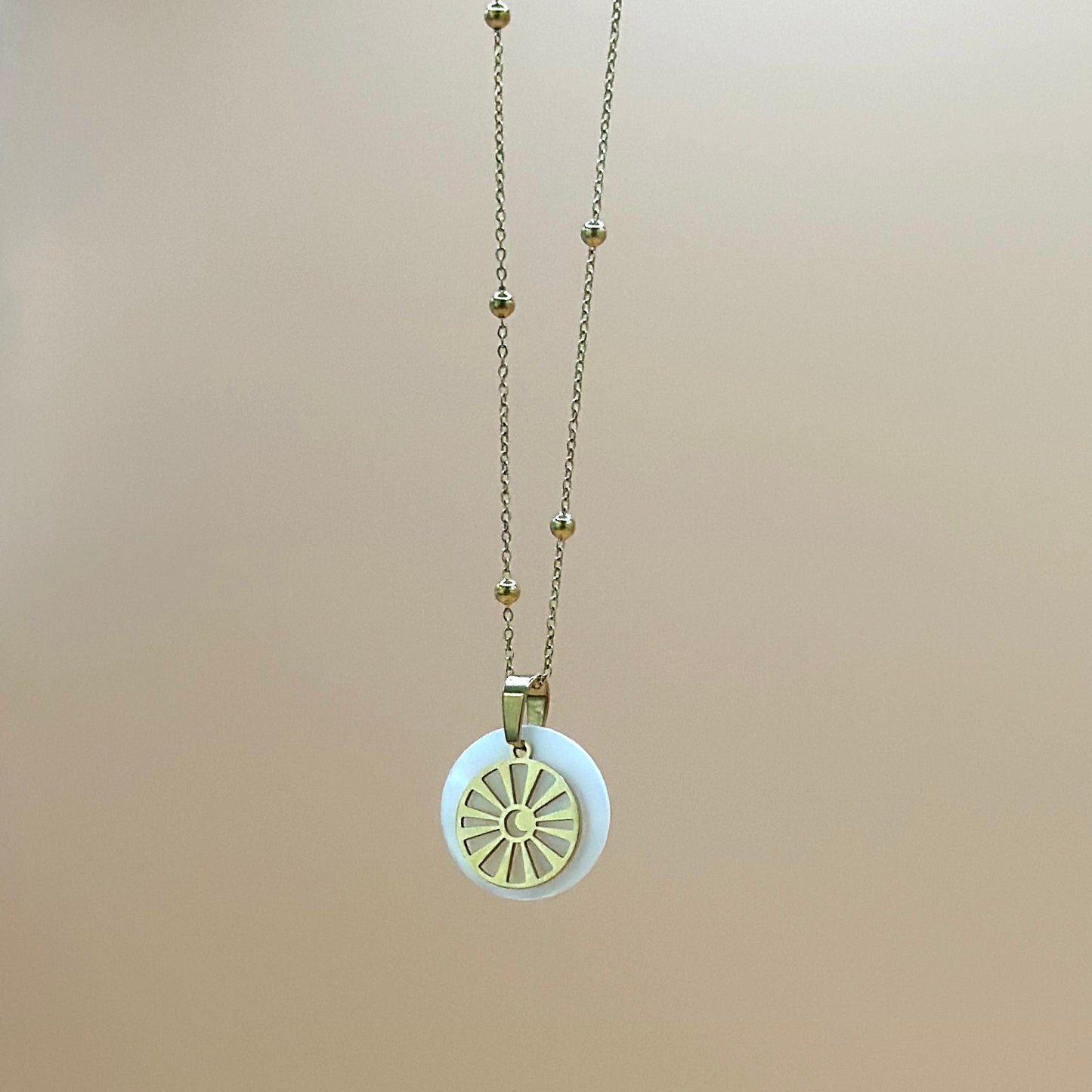 Goldfarbene Halskette auf lachsfarbenem Hintergrund. Die Kette hat kleinen Kugeln als Details und der Anhänger ist rund und zeigt eine Sonne in deren Zentrum ein Mond zu sehen ist. Der Anhänger liegt auf einem Perlmuttplättchen