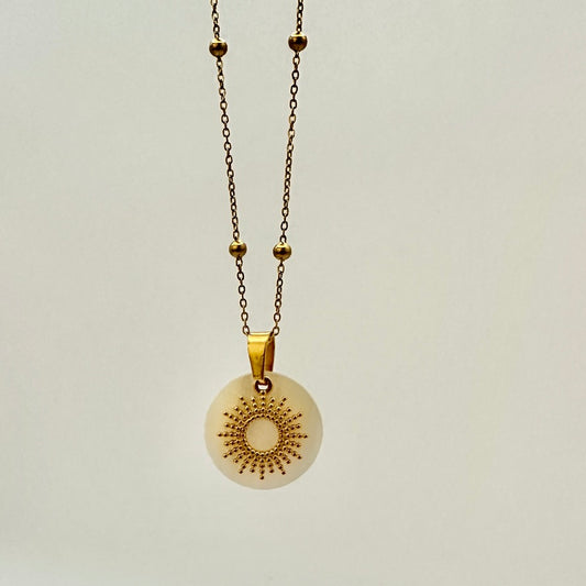 Goldfarbene Halskette auf weißem Hintergrund. Die Kette hat kleinen Kugeln als Details und der Anhänger ist eine Sonne, deren Strahlen kleine, aneinander gereihte Kugeln sind. Der Anhänger liegt auf einem Perlmuttplättchen.