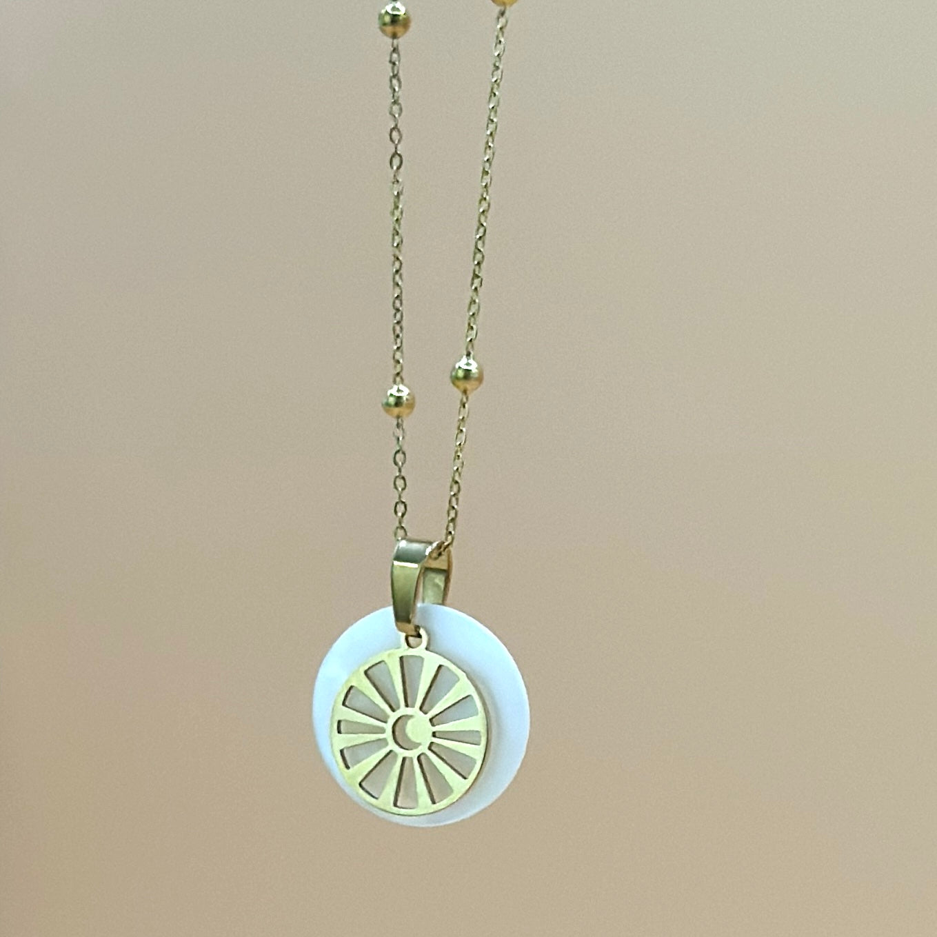 Goldfarbene Halskette auf lachsfarbenem Hintergrund. Die Kette hat kleinen Kugeln als Details und der Anhänger ist rund und zeigt eine Sonne in deren Zentrum ein Mond zu sehen ist. Der Anhänger liegt auf einem Perlmuttplättchen