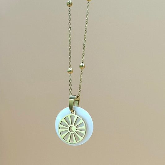 Goldfarbene Halskette auf lachsfarbenem Hintergrund. Die Kette hat kleinen Kugeln als Details und der Anhänger ist rund und zeigt eine Sonne in deren Zentrum ein Mond zu sehen ist. Der Anhänger liegt auf einem Perlmuttplättchen