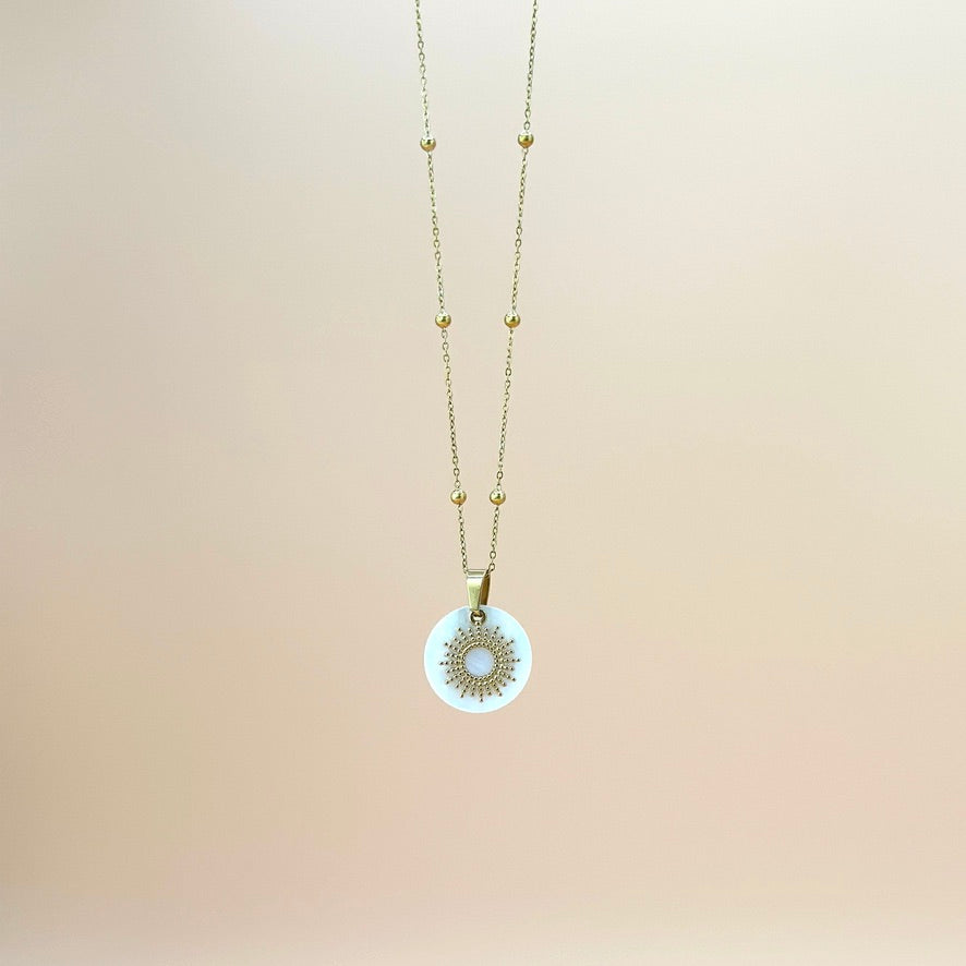 Goldfarbene Halskette auf lachsfarbenem Hintergrund. Die Kette hat kleinen Kugeln als Details und der Anhänger ist eine Sonne, deren Strahlen kleine, aneinander gereihte Kugeln sind. Der Anhänger liegt auf einem Perlmuttplättchen.