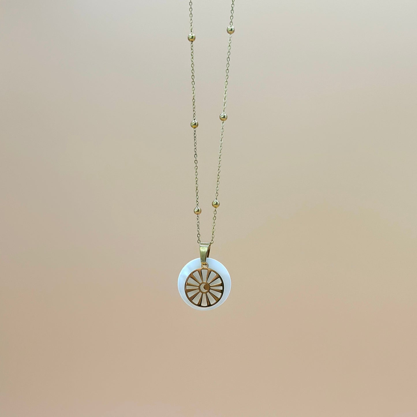 Goldfarbene Halskette auf lachsfarbenem Hintergrund. Die Kette hat kleinen Kugeln als Details und der Anhänger ist rund und zeigt eine Sonne in deren Zentrum ein Mond zu sehen ist. Der Anhänger liegt auf einem Perlmuttplättchen