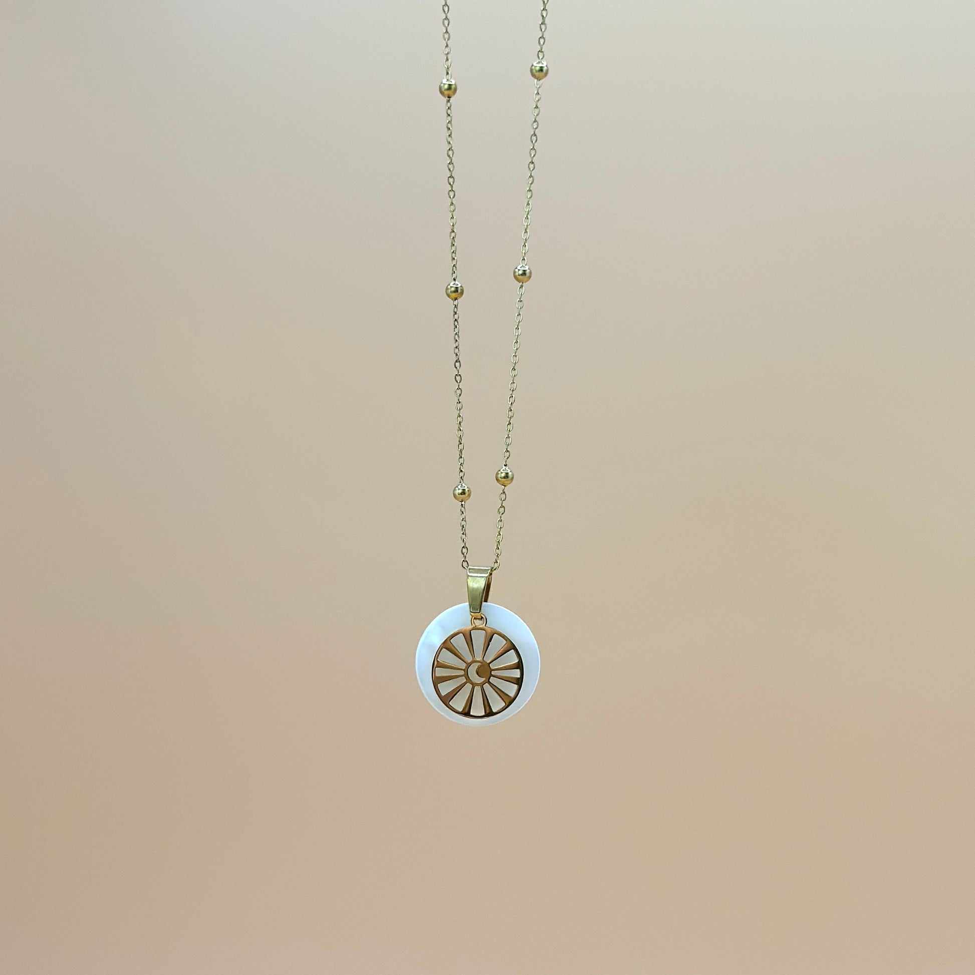 Goldfarbene Halskette auf lachsfarbenem Hintergrund. Die Kette hat kleinen Kugeln als Details und der Anhänger ist rund und zeigt eine Sonne in deren Zentrum ein Mond zu sehen ist. Der Anhänger liegt auf einem Perlmuttplättchen