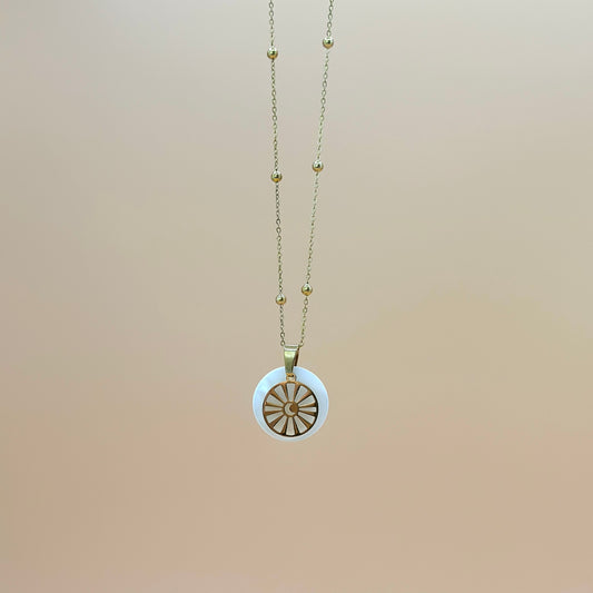 Goldfarbene Halskette auf lachsfarbenem Hintergrund. Die Kette hat kleinen Kugeln als Details und der Anhänger ist rund und zeigt eine Sonne in deren Zentrum ein Mond zu sehen ist. Der Anhänger liegt auf einem Perlmuttplättchen