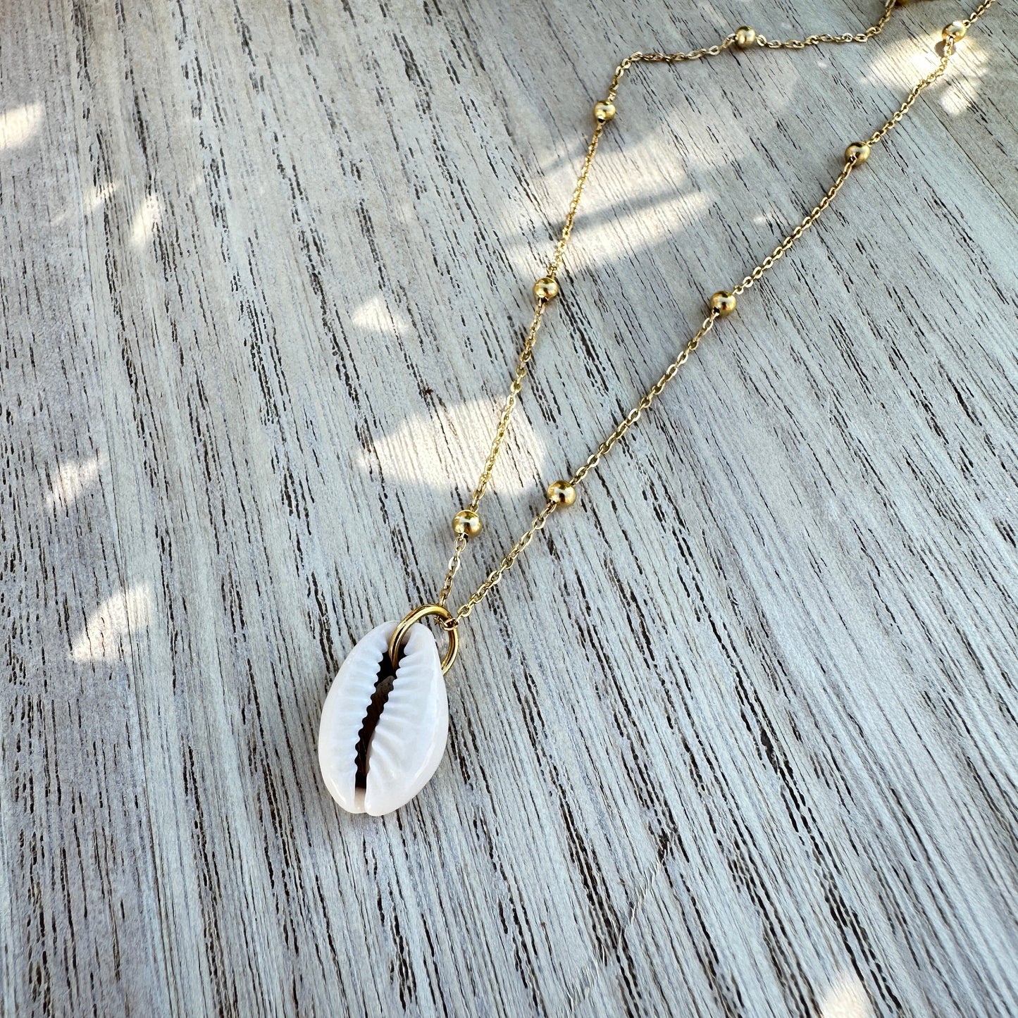 Weiße Kaurimuschel-Halskette mit goldener Edelstahlkette flachliegend auf Holztisch.